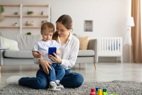 Mother sitting on a living room floor with her young child while looking at a smartphone, representing parenting responsibilities, child wellbeing, and financial planning after separation or divorce.